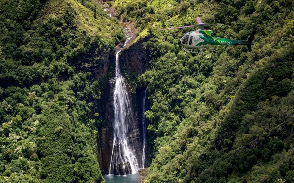A helicopter flies low over the ridgelines and sea cliffs of Kauai